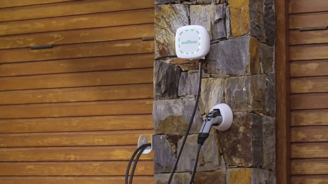 Fehér Wallbox Pulsar Plus fali töltő kő- és fa burkolatú házfalon, a töltőkábel a falra szerelt tartóban