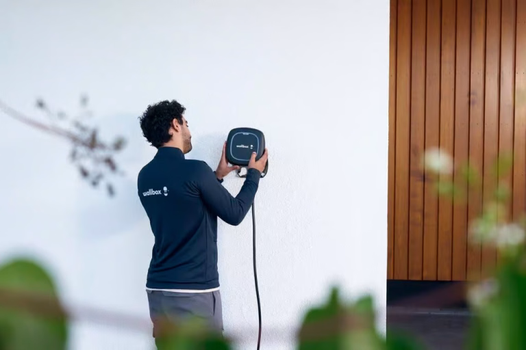Wallbox-feliratú munkaruhás szerelő fekete fali töltőt szerel fel egy fehér homlokzatra