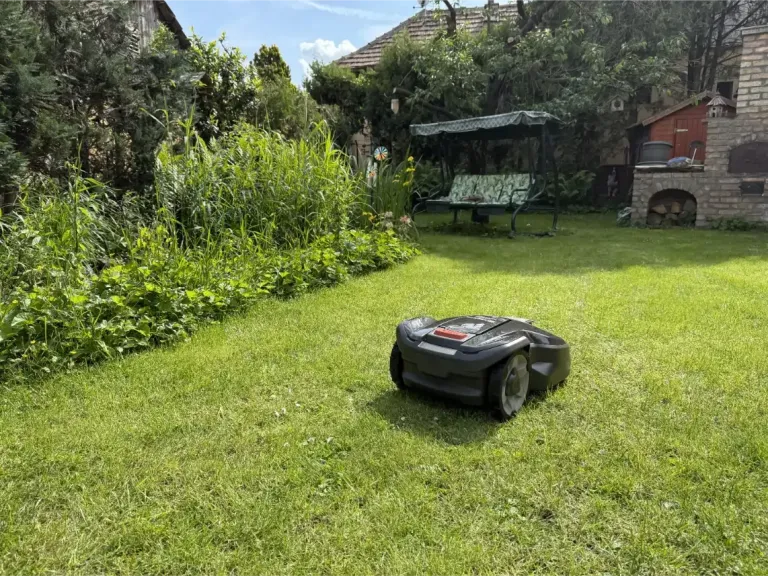 Husqvarna robotfűnyíró frissen nyírt pázsiton egy napos kertben, háttérben kerti hinta és téglából rakott kemence