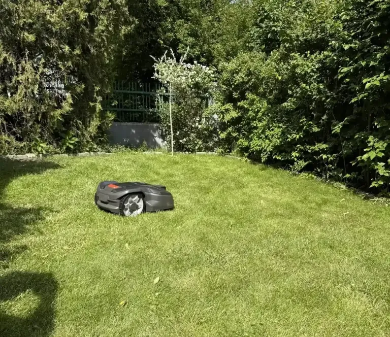 Szürke Husqvarna robotfűnyíró a nyírt gyepen, sűrű sövénnyel és fákkal körülvett napos kertben