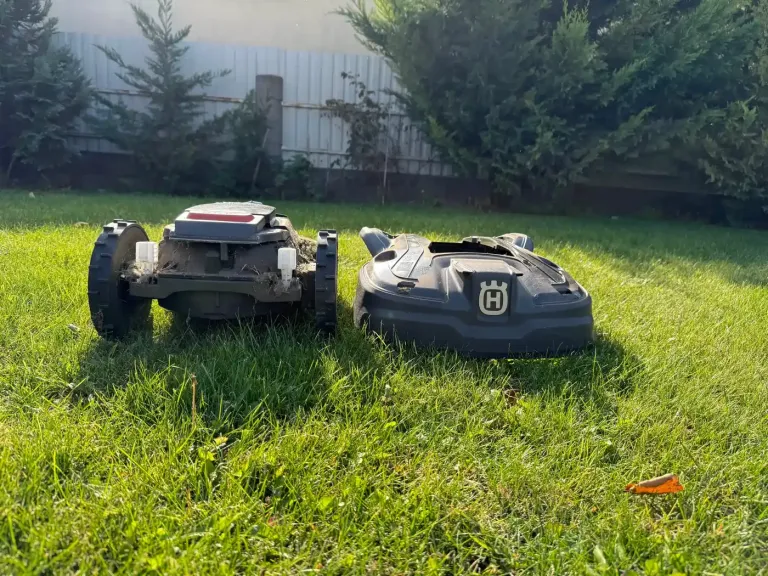 Szétszedett Husqvarna robotfűnyíró a gyepen: a fűnyesedékes alváz és mellette a levett burkolat, esti napfényben