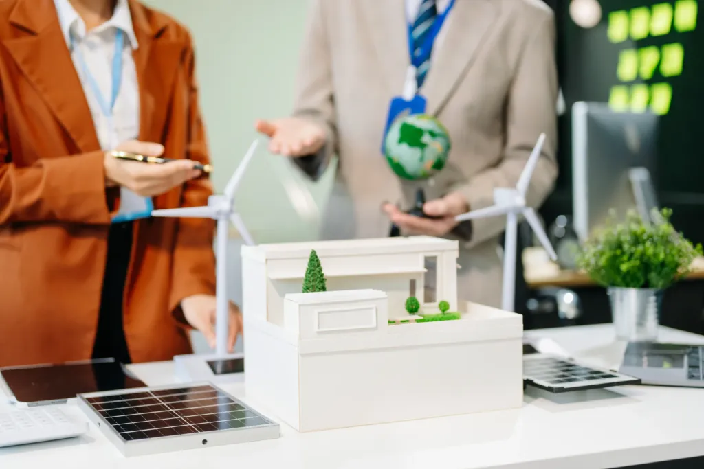 Üzleti megbeszélés fehér épületmakett, napelem- és szélturbina-modellek körül egy irodai asztalnál